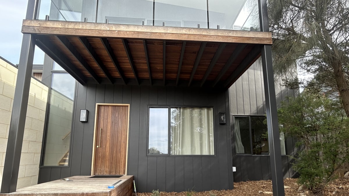 Entry elevation of a dark-clad house with a timber-lined entry canopy, a timber front door and a glass balustrade above, Mornington Peninsula.